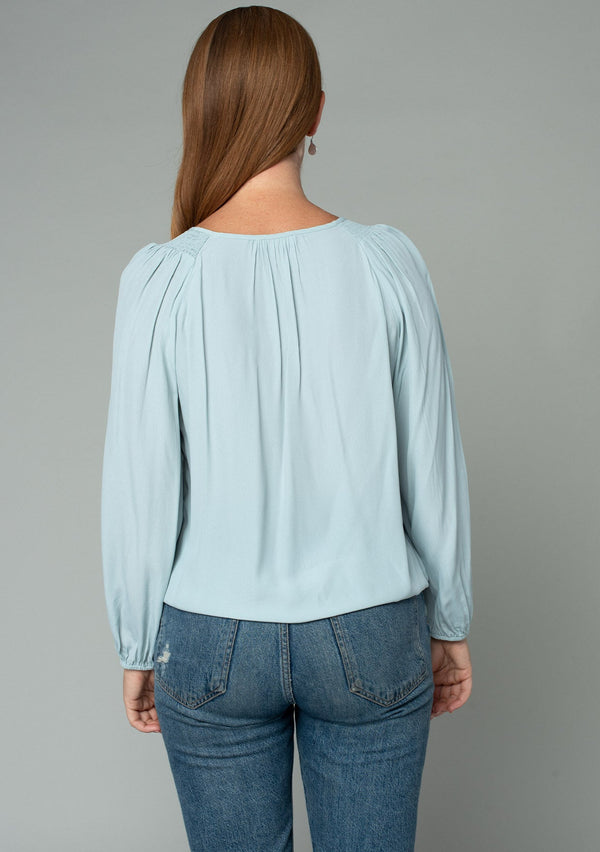 [Color: Sky] A back facing image of a red headed model wearing a soft and silky light blue bohemian blouse with voluminous long sleeves, a plunging surplice v neckline, and an elastic front waist.