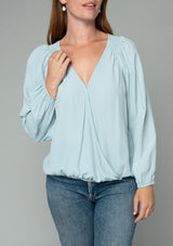 [Color: Sky] A close up front facing image of a red headed model wearing a soft and silky light blue bohemian blouse with voluminous long sleeves, a plunging surplice v neckline, and an elastic front waist.