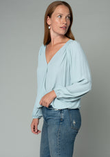 [Color: Sky] A side facing image of a red headed model wearing a soft and silky light blue bohemian blouse with voluminous long sleeves, a plunging surplice v neckline, and an elastic front waist.