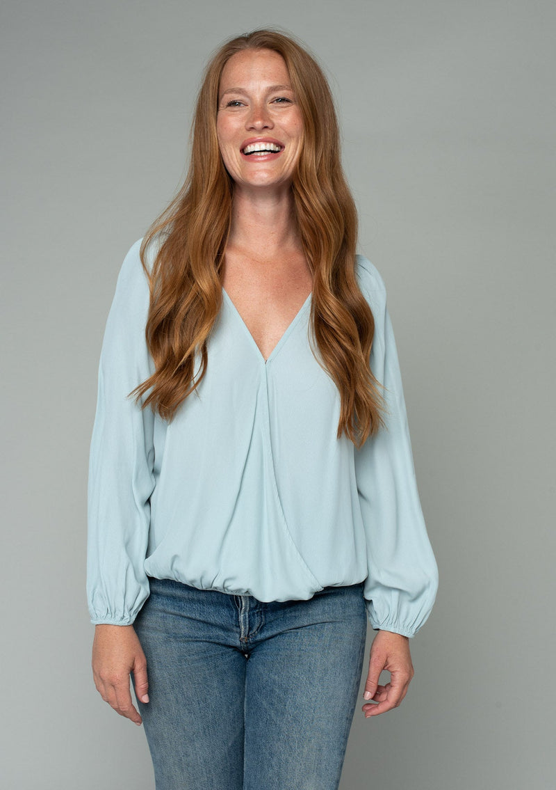 [Color: Sky] A front facing image of a red headed model wearing a soft and silky light blue bohemian blouse with voluminous long sleeves, a plunging surplice v neckline, and an elastic front waist. The model has long wavy red hair. 