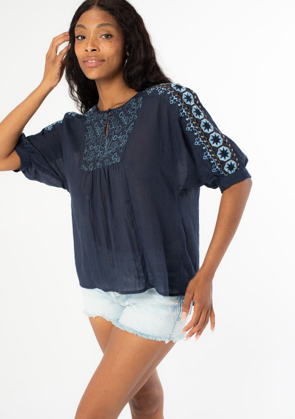 [Color: Navy] An angled front facing image of a black model wearing a navy blue cotton bohemian top with half length dolman sleeves and a contrast light blue embroidered detail along the sleeve and front bib. 