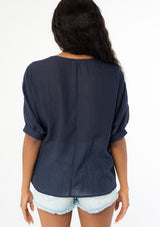[Color: Navy] A back facing image of a black model wearing a navy blue cotton bohemian top with half length dolman sleeves and a contrast light blue embroidered detail along the sleeve and front bib. 