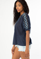 [Color: Navy] A side facing image of a black model wearing a navy blue cotton bohemian top with half length dolman sleeves and a contrast light blue embroidered detail along the sleeve and front bib. 