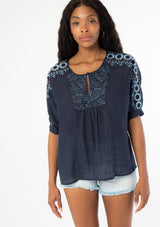 [Color: Navy] A front facing image of a black model wearing a navy blue cotton bohemian top with half length dolman sleeves and a contrast light blue embroidered detail along the sleeve and front bib. 