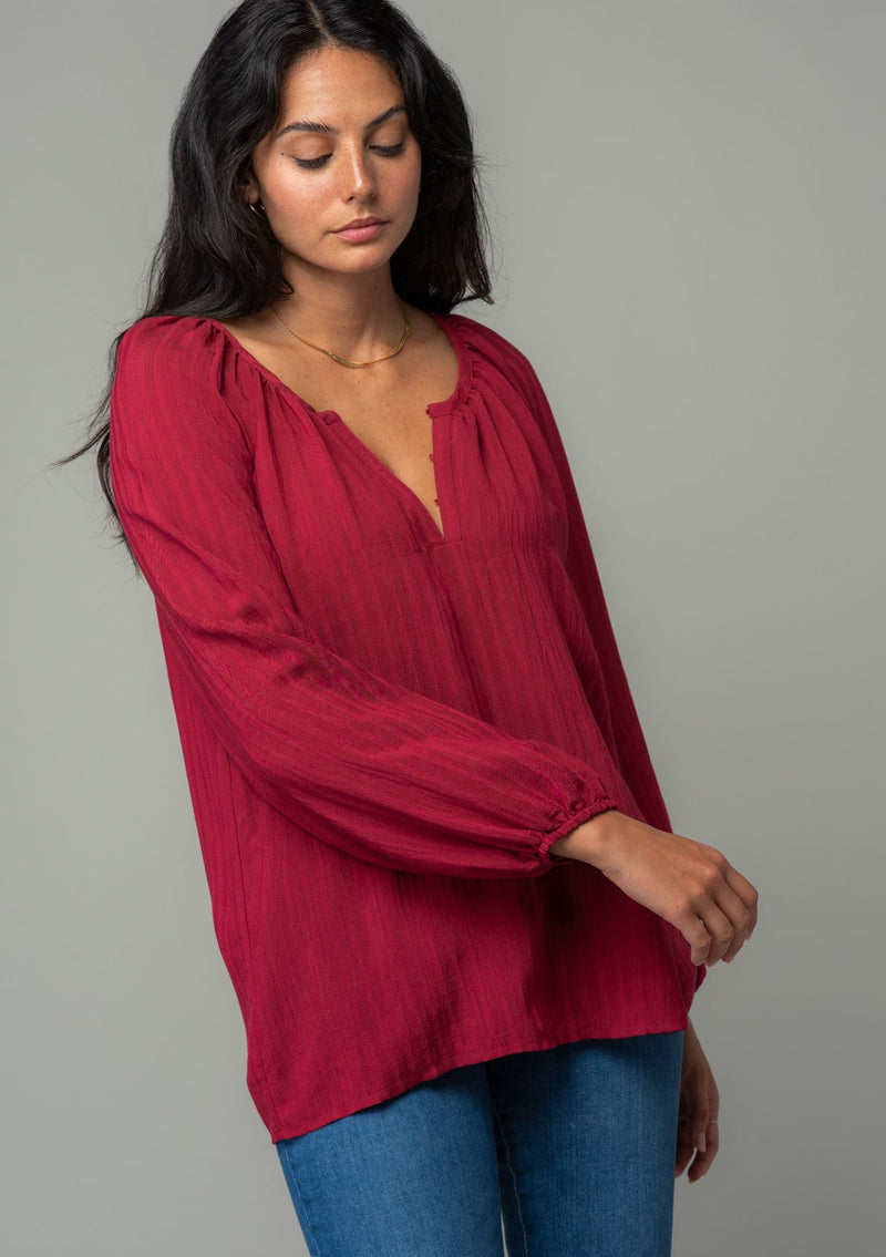 [Color: Burgundy] A front facing image of a brunette model wearing a burgundy red flowy bohemian peasant top in a textured shadow stripe. With voluminous long sleeves, a split v neckline with hook and eye closure, and a round neckline. 