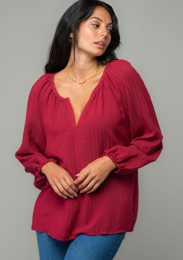 [Color: Burgundy] A half body front facing image of a brunette model wearing a burgundy red flowy bohemian peasant top in a textured shadow stripe. With voluminous long sleeves, a split v neckline with hook and eye closure, and a round neckline. 