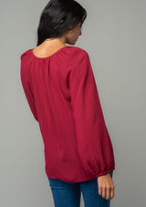 [Color: Burgundy] A back facing image of a brunette model wearing a burgundy red flowy bohemian peasant top in a textured shadow stripe. With voluminous long sleeves, a split v neckline with hook and eye closure, and a round neckline. 