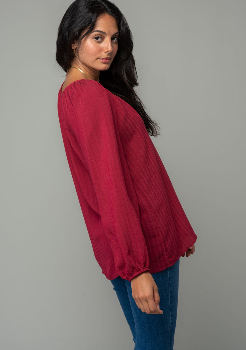 [Color: Burgundy] A side facing image of a brunette model wearing a burgundy red flowy bohemian peasant top in a textured shadow stripe. With voluminous long sleeves, a split v neckline with hook and eye closure, and a round neckline. 