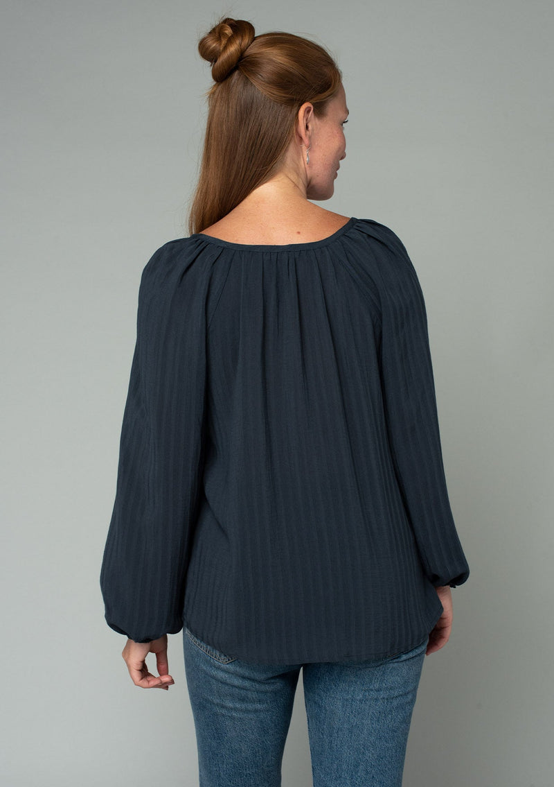 [Color: Charcoal] A back facing image of a red headed model wearing a dark grey flowy bohemian peasant top in a textured shadow stripe. With voluminous long sleeves, a split v neckline with hook and eye closure, and a round neckline.