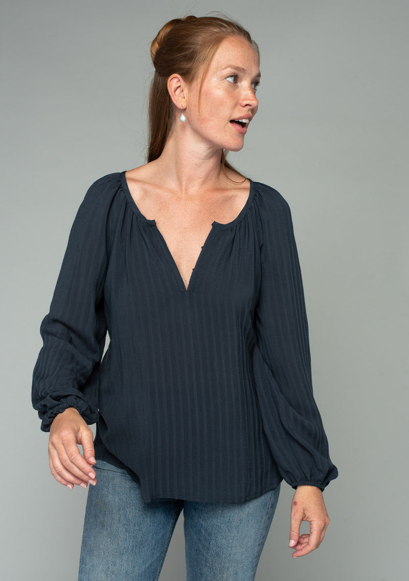 [Color: Charcoal] A front facing image of a red headed model wearing a dark grey flowy bohemian peasant top in a textured shadow stripe. With voluminous long sleeves, a split v neckline with hook and eye closure, and a round neckline.