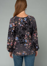 [Color: Grey/Dusty Blue] A back facing image of a red headed model wearing a classic bohemian peasant blouse in a grey and blue floral print. With long voluminous raglan sleeves and a front placket with a hook and eye closure. 