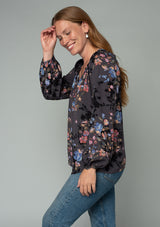 [Color: Grey/Dusty Blue] A side facing image of a red headed model wearing a classic bohemian peasant blouse in a grey and blue floral print. With long voluminous raglan sleeves and a front placket with a hook and eye closure. 