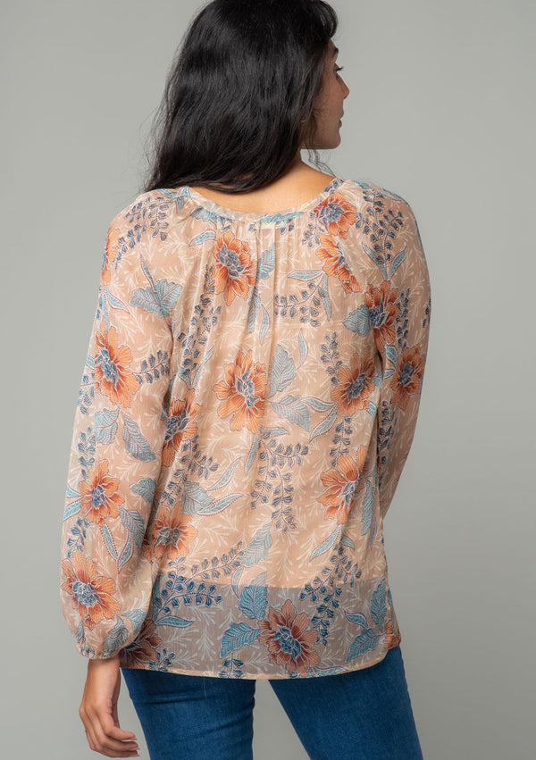 [Color: Coral/Blue] A back facing image of a brunette model wearing a sheer chiffon bohemian blouse in a coral and blue floral print. With long sleeves, a relaxed flowy fit, and a hook and eye closure neckline. 