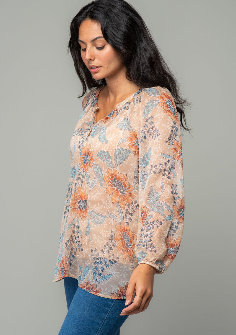 [Color: Coral/Blue] A side facing image of a brunette model wearing a sheer chiffon bohemian blouse in a coral and blue floral print. With long sleeves, a relaxed flowy fit, and a hook and eye closure neckline. 