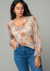 [Color: Coral/Blue] A front facing image of a brunette model wearing a sheer chiffon bohemian blouse in a coral and blue floral print. With long sleeves, a relaxed flowy fit, and a hook and eye closure neckline. The model is wearing the blouse tucked into denim. 