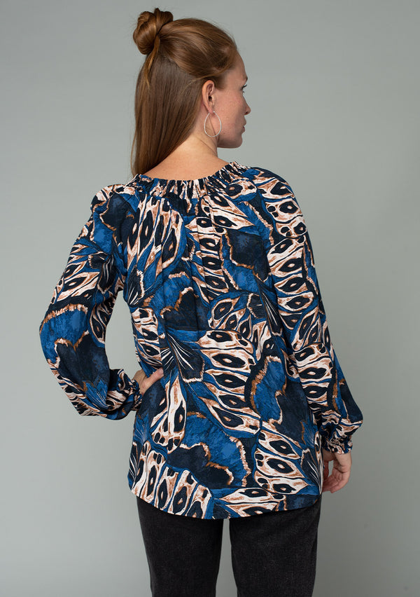 [Color: Cobalt/Tan] A back facing image of a red headed model wearing a bohemian peasant top in a navy blue and tan abstract butterfly wing print. With voluminous long sleeves, a smocked round neckline with tassel ties, and a flowy relaxed fit. 