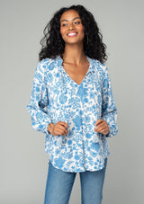[Color: Cream/Dusty Blue] A half body front facing image of a brunette model wearing a classic bohemian peasant top in a white and blue floral print. With voluminous long sleeves, a smocked neckline, tassel neck ties, and a loose flowy fit. 