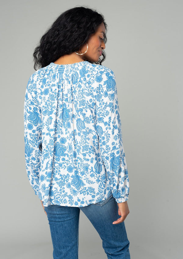 [Color: Cream/Dusty Blue] A back facing image of a brunette model wearing a classic bohemian peasant top in a white and blue floral print. With voluminous long sleeves, a smocked neckline, tassel neck ties, and a loose flowy fit. 