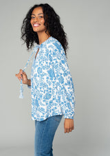 [Color: Cream/Dusty Blue] A side facing image of a brunette model wearing a classic bohemian peasant top in a white and blue floral print. With voluminous long sleeves, a smocked neckline, tassel neck ties, and a loose flowy fit. 