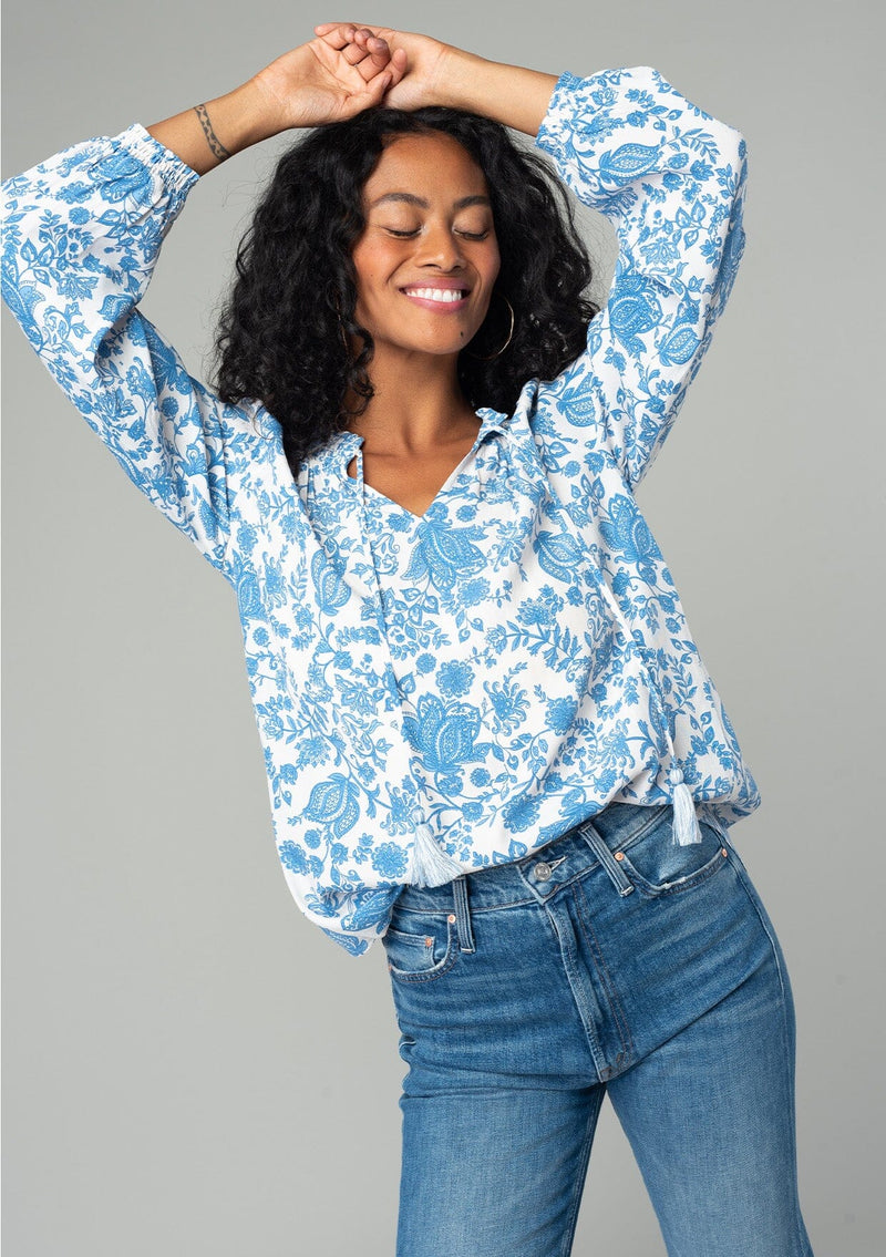 [Color: Cream/Dusty Blue] A front facing image of a brunette model wearing a classic bohemian peasant top in a white and blue floral print. With voluminous long sleeves, a smocked neckline, tassel neck ties, and a loose flowy fit. 