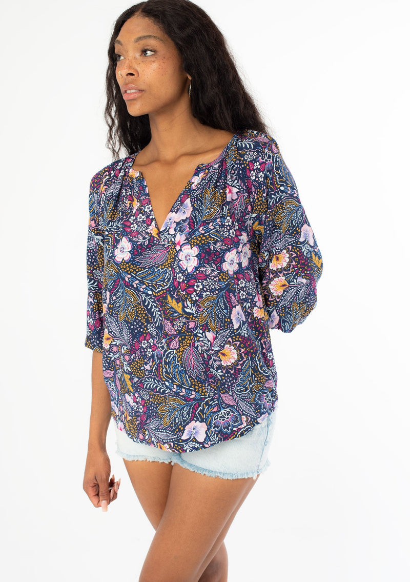 [Color: Navy/Berry] A semi side facing image of a black model with long dark wavy hair wearing a navy blue and berry purple floral print top. With three quarter length sleeves and a v neckline. 