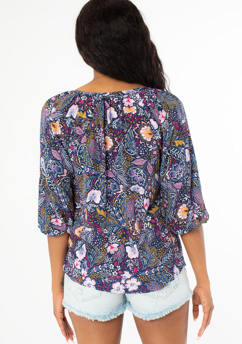 [Color: Navy/Berry] A back facing image of a black model with long dark wavy hair wearing a navy blue and berry purple floral print top. With three quarter length sleeves and a v neckline. 