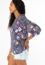[Color: Navy/Berry] A side facing image of a black model with long dark wavy hair wearing a navy blue and berry purple floral print top. With three quarter length sleeves and a v neckline. 