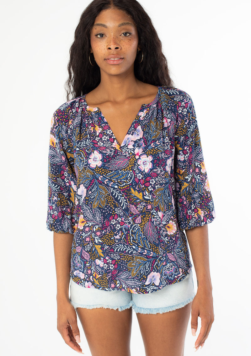[Color: Navy/Berry] A front facing image of a black model with long dark wavy hair wearing a navy blue and berry purple floral print top. With three quarter length sleeves and a v neckline. 