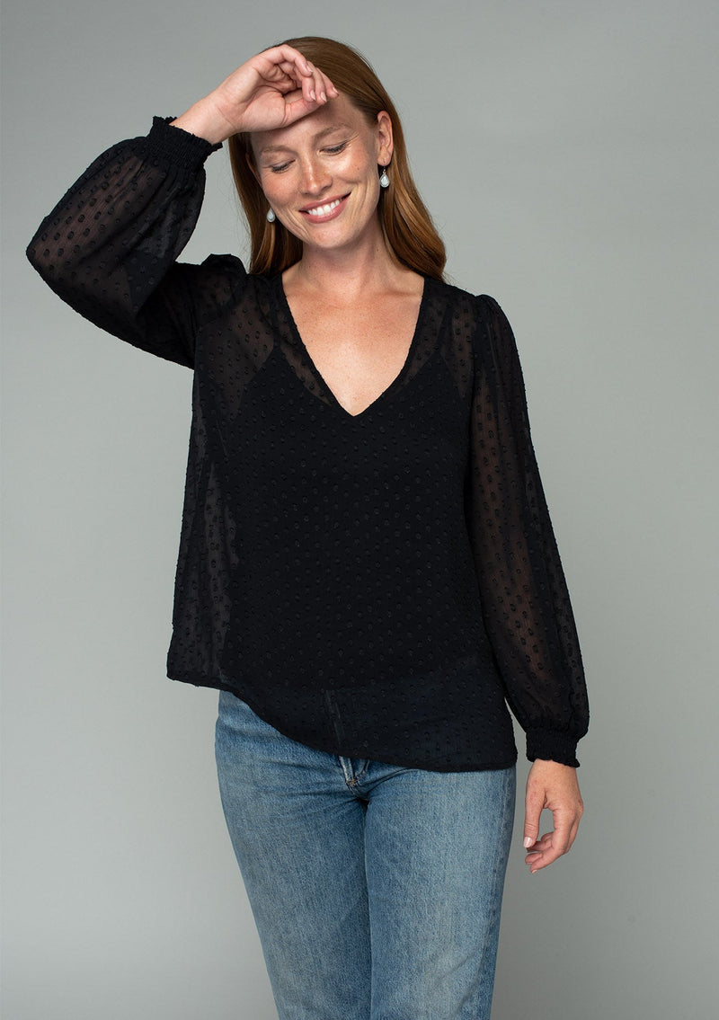 [Color: Black] A front facing image of a red headed model wearing a black bohemian blouse in a light catching clip dot sheer chiffon. With voluminous long sleeves, a v neckline, and a relaxed flowy fit. 