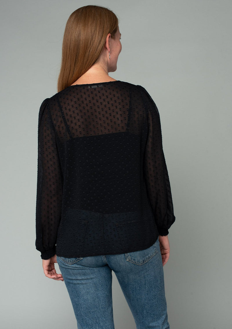 [Color: Black] A back facing image of a red headed model wearing a black bohemian blouse in a light catching clip dot sheer chiffon. With voluminous long sleeves, a v neckline, and a relaxed flowy fit. 