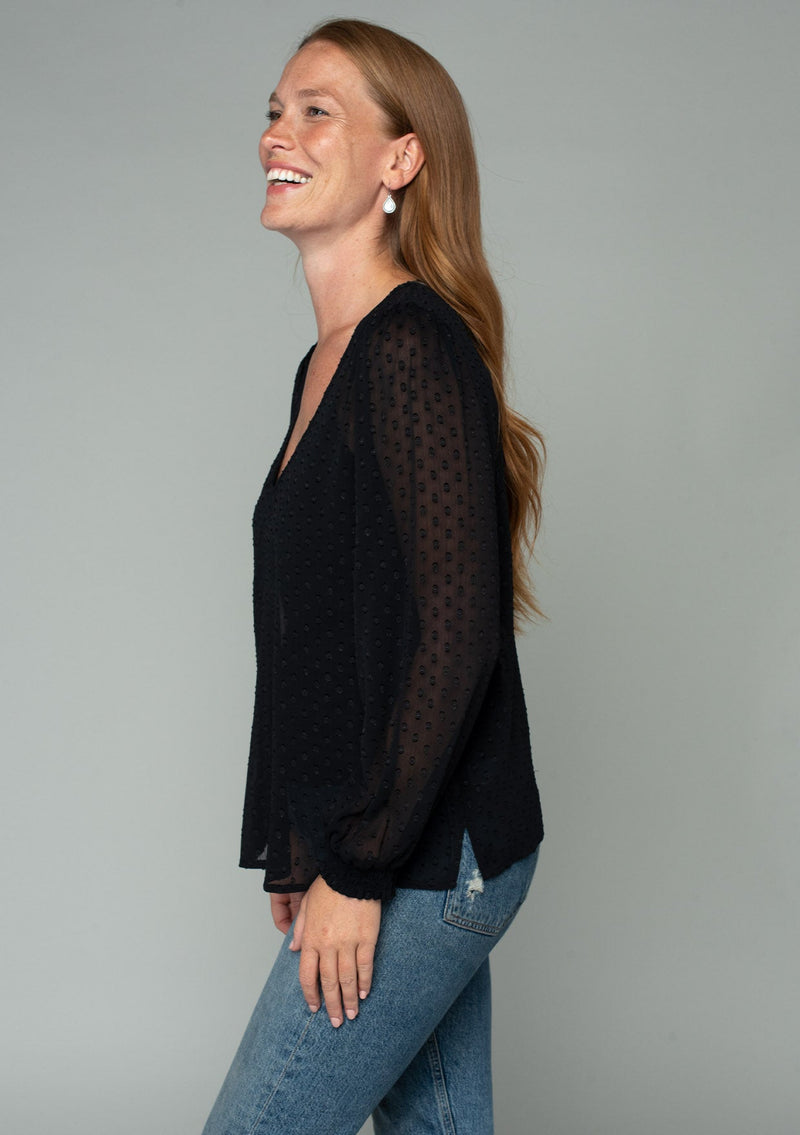 [Color: Black] A side facing image of a red headed model wearing a black bohemian blouse in a light catching clip dot sheer chiffon. With voluminous long sleeves, a v neckline, and a relaxed flowy fit. 