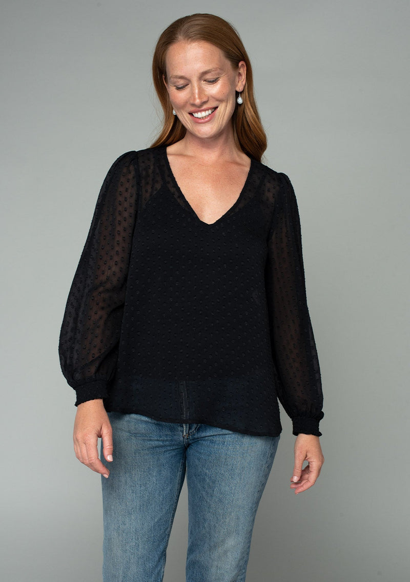 [Color: Black] A front facing image of a red headed model wearing a black bohemian blouse in a light catching clip dot sheer chiffon. With voluminous long sleeves, a v neckline, and a relaxed flowy fit. 