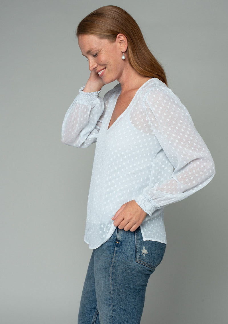 [Color: Ice Blue] A side facing image of a red headed model wearing a light blue bohemian blouse in a light catching clip dot sheer chiffon. With voluminous long sleeves, a v neckline, and a relaxed flowy fit. 