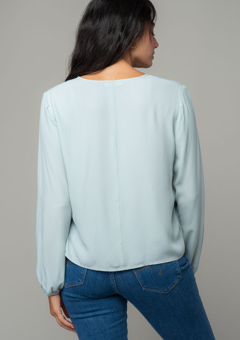 [Color: Sky] A back facing image of a brunette model wearing a soft and silky light blue crepe long sleeve top with a tie front detail. 