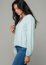 [Color: Sky] A side facing image of a brunette model wearing a soft and silky light blue crepe long sleeve top with a tie front detail. 
