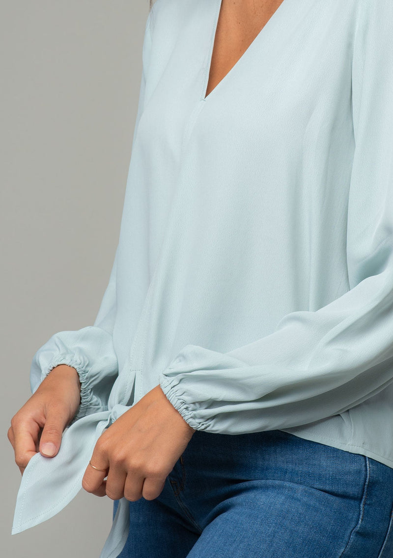 [Color: Sky] A close up side facing image of a brunette model wearing a soft and silky light blue crepe long sleeve top with a tie front detail. 