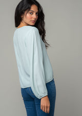 [Color: Sky] A side facing image of a brunette model wearing a soft and silky light blue crepe long sleeve top with a tie front detail. The model is looking over her shoulder. 