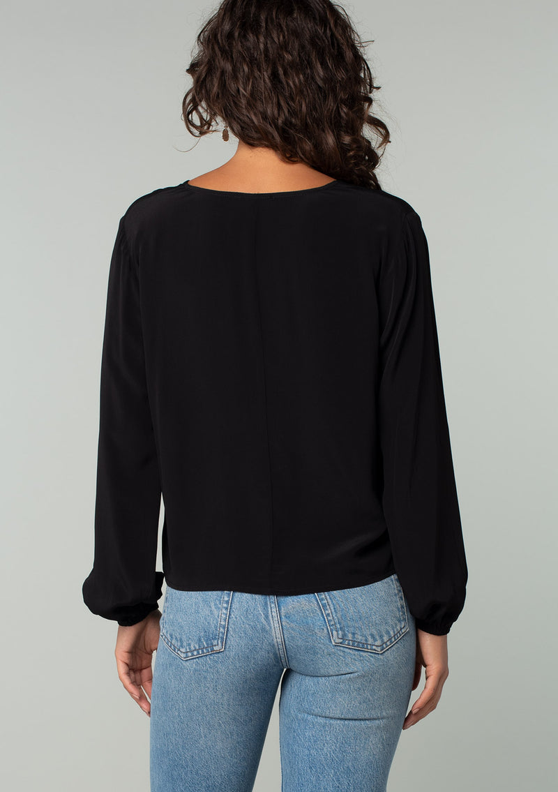 [Color: Black] A back facing image of a brunette model wearing a soft and silky black crepe long sleeve top with a tie front detail.