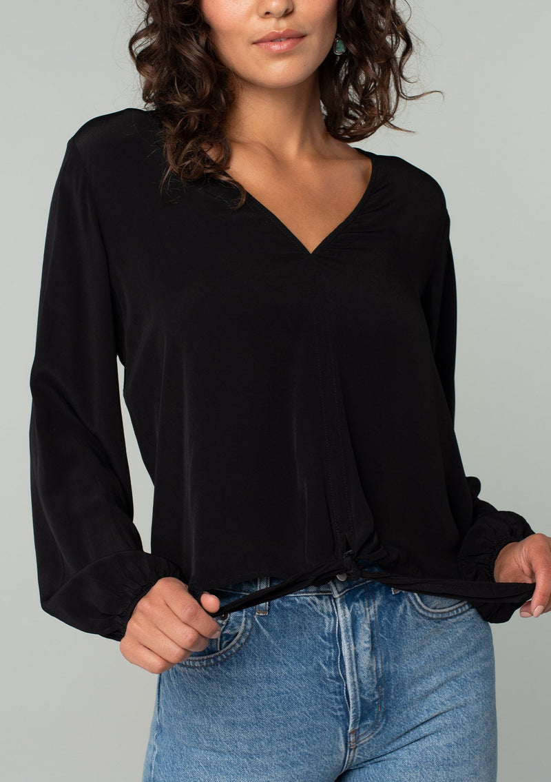 [Color: Black] A close up front facing image of a brunette model wearing a soft and silky black crepe long sleeve top with a tie front detail.