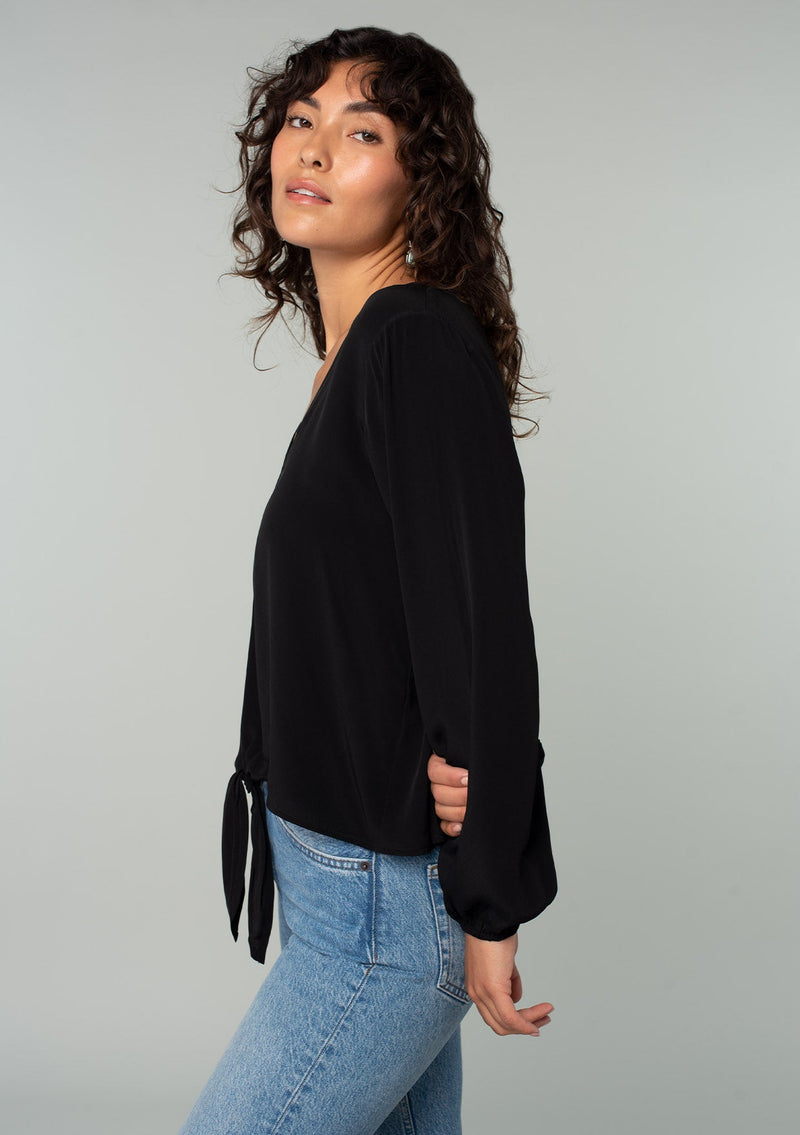 [Color: Black] A side facing image of a brunette model wearing a soft and silky black crepe long sleeve top with a tie front detail.