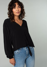 [Color: Black] A front facing image of a brunette model wearing a soft and silky black crepe long sleeve top with a tie front detail.