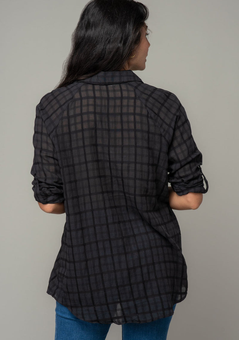 [Color: Black] A back facing image of a brunette model wearing a soft black cotton button front shirt in a textured gingham fabric. With long rolled sleeves, a button front, and a collared neckline.