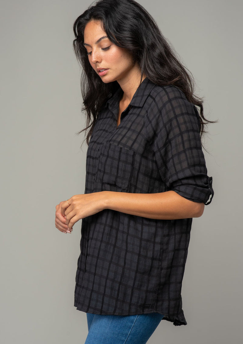 [Color: Black] A side facing image of a brunette model wearing a soft black cotton button front shirt in a textured gingham fabric. With long rolled sleeves, a button front, and a collared neckline.