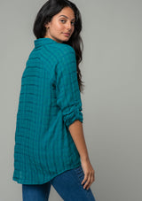 [Color: Teal] A back facing image of a brunette model wearing a soft teal cotton button front shirt in a textured gingham fabric. With long rolled sleeves, a button front, and a collared neckline. The model is looking over her shoulder. 