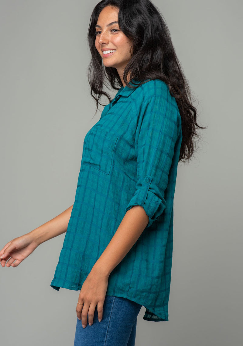 [Color: Teal] A side facing image of a brunette model wearing a soft teal cotton button front shirt in a textured gingham fabric. With long rolled sleeves, a button front, and a collared neckline.