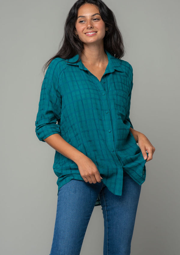 [Color: Teal] A front facing image of a brunette model wearing a soft teal cotton button front shirt in a textured gingham fabric. With long rolled sleeves, a button front, and a collared neckline.