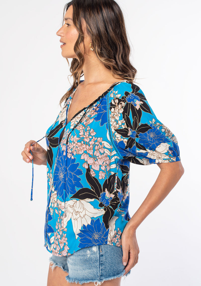 [Color: Turquoise/Black] A woman wearing a flowy bohemian top in a blue and black large floral print. With half length puff sleeves and a button front. 