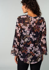 [Color: Black/Dusty Rose] A back facing image of a brunette model wearing a sheer chiffon bohemian blouse in a black and rose pink floral print. With long sleeves, ruffled wrist cuffs, and a split v neckline. 
