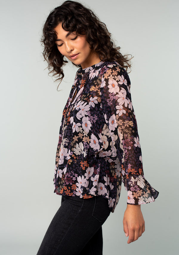 [Color: Black/Dusty Rose] A side facing image of a brunette model wearing a sheer chiffon bohemian blouse in a black and rose pink floral print. With long sleeves, ruffled wrist cuffs, and a split v neckline. 