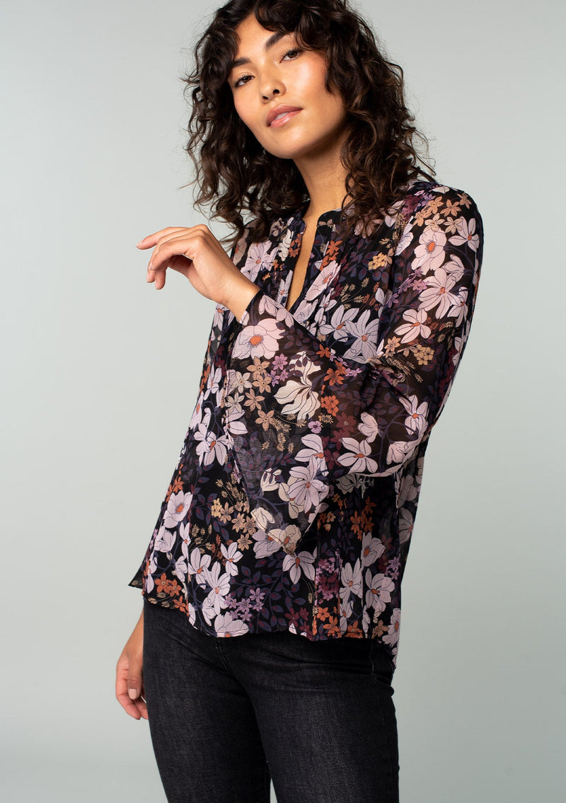[Color: Black/Dusty Rose] An angled front facing image of a brunette model wearing a sheer chiffon bohemian blouse in a black and rose pink floral print. With long sleeves, ruffled wrist cuffs, and a split v neckline. 